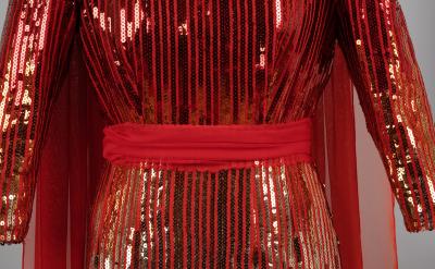 Close-up of a garment with vertical red and gold sequin stripes and sheer red fabric accents; features long sleeves and a solid red waistband or sash at the midsection.