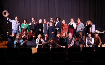 A group of about 25 adults, some in costume, pose and smile on a stage in front of a black curtain. Some are sitting, some standing, with one person raising a hat and others making joyful gestures.