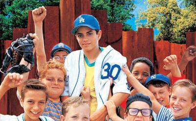 A group of smiling boys in baseball uniforms and casual clothes pose together, raising their fists in excitement. The title "The Sandlot" appears in large yellow letters above them against a bright blue sky.