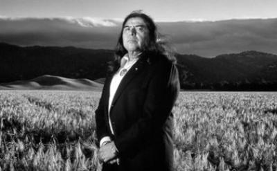 A man with long hair, dressed in a suit, stands in a field of tall grass with mountains and dramatic clouds in the background. The image is in black and white.