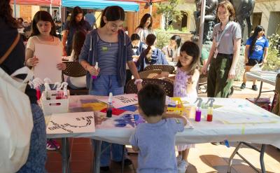 Children and adults gather around a table under a red canopy, engaging in arts and crafts activities with paints and paper at an outdoor event. People chat and work together in a lively, sunny environment.