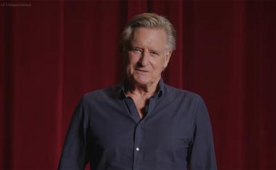A man with gray hair wearing a navy shirt stands in front of red theater curtains, looking at the camera and smiling gently.