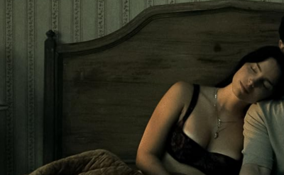 A woman sleeps with her head on a man's shoulder as they sit on a bed in a dimly lit room with patterned wallpaper and a lamp on the nightstand.
