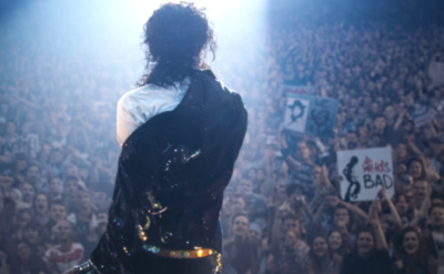 A performer stands on stage facing a large, cheering crowd with spotlights shining down. Some audience members hold up signs, including one that says "Bad.