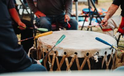 Four people sit around a large traditional drum, each holding a colored mallet and striking the drum together. The drum has a taut, laced surface and is played outdoors.