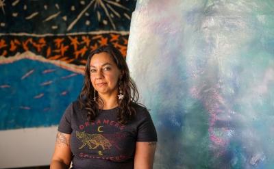 A woman with long brown hair and tattoos sits in front of colorful, abstract artwork. She wears a “Dreamers & Schemers” t-shirt and star earrings, smiling gently at the camera.