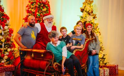 A family and an elderly woman pose for a photo with Santa Claus, who is seated in a red sleigh between two decorated Christmas trees.