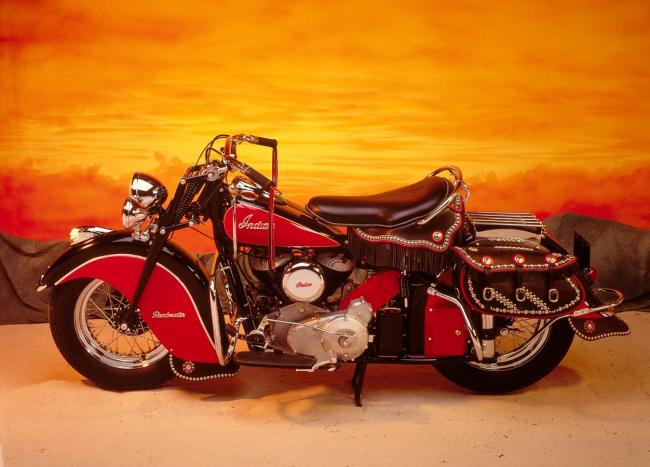A motorcycle with the word Indian painted upon it in front of a backdrop painted like the sky