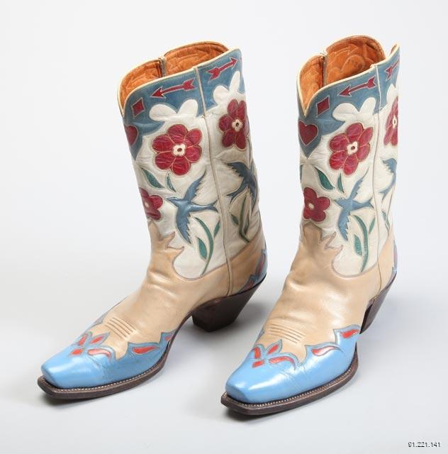 Pair of leather cowboy boots stand against a grey background, their toes angled slightly to the left. The boots are adorned with a blue, red, and cream design. Sky blue birds open their wings in flight between blades of green grass and red flowers. Near the top of the shoe there is a strip of sky-blue leather with red arrows and a large red heart. The boots have a dark-brown heel.