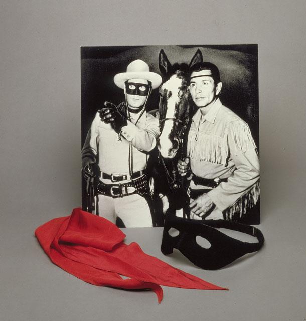 A red neck scarf, black mask, and black and white photograph rest against a grey-blue background. The photograph depicts actor Clayton Moore in his Lone Ranger costume including the black domino mask and neck scarf. Jay Silverheels, dressed as his Long Ranger character Tonto stands to Moore’s right and holds the reins on Scout, Tonto’s horse on the show. Scout leans his head forward between Silverheels and Moore as Moore points forward. In front of the black and white photograph, the red neck scarf is laid 
