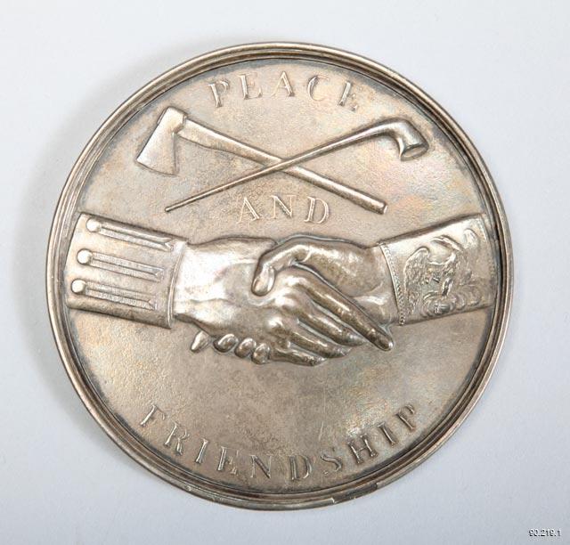 Silver medal coin with a raised design. Design depicts two hands clasped in the center in a handshake. The left arm has a sleeve designed with three service stripes, set with buttons. The right arm has a Colonel Eagle on its sleeve. Above the joined hands is a crossed pipe and hatchet. In all-caps and a serif font the words “Peace and Friendship” are spaced evenly across the coin face; “Peace” sits above the crossed tools, “And” between the tools and the hands, and “Friendship” below the joined hands, curve
