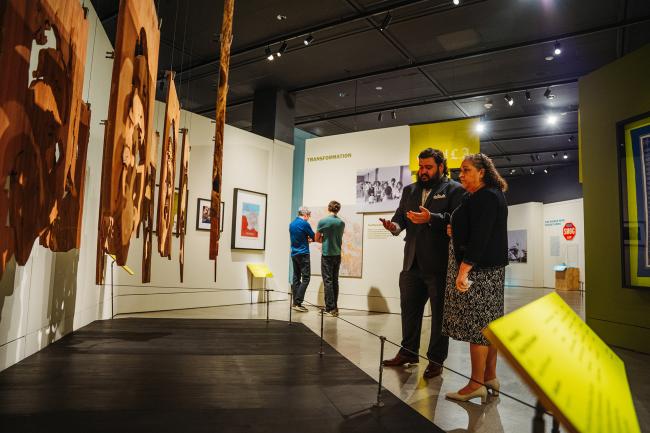 Two people stand in a museum exhibit, discussing wooden hanging art pieces. Other visitors observe displays in the background. The space is well-lit with informational signs and photos on the walls.