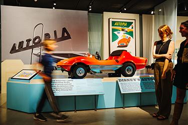 A red Disney Autopia car is displayed in a museum exhibit, with large Autopia signs and posters in the background. Two people stand nearby while another person walks past, slightly blurred.