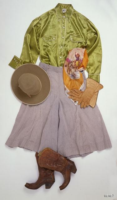 A Western-style ensemble is laid flat in a composite, as if someone had laid out the outfit before wearing it. The outfit consists of a chartreuse satin button-down shirt with two breast pockets. The shirt has round, white buttons and crisp white edging along the collar, front placket, and breast flap pockets. Laying atop the bottom of the western shirt, a faded purple-grey split-skirt is splayed out wide. Placed casually along the left hip of the shirt is an orange and pastel-pink silk scarf with a white, 