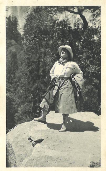 woman standing in the wilderness at the edge of a rock, she wears old style outdoors outfit with hat and boots