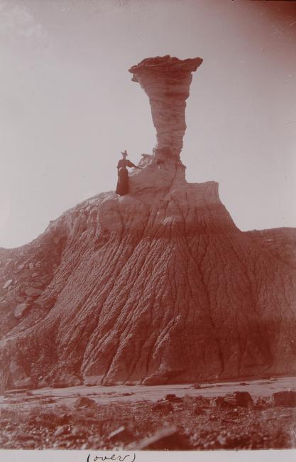 woman in 19th century clothing stands on steep hill next to rock formation