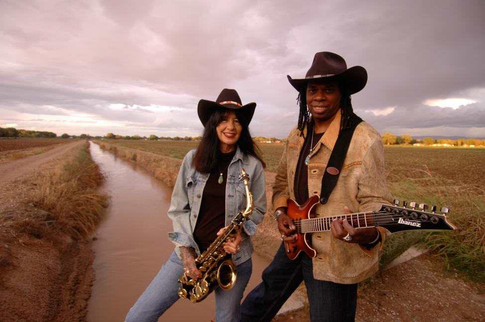 two women musicians in a field at an acquaduct both wear brown cowboy hats one holds a saxophone and the other holds a guitar
