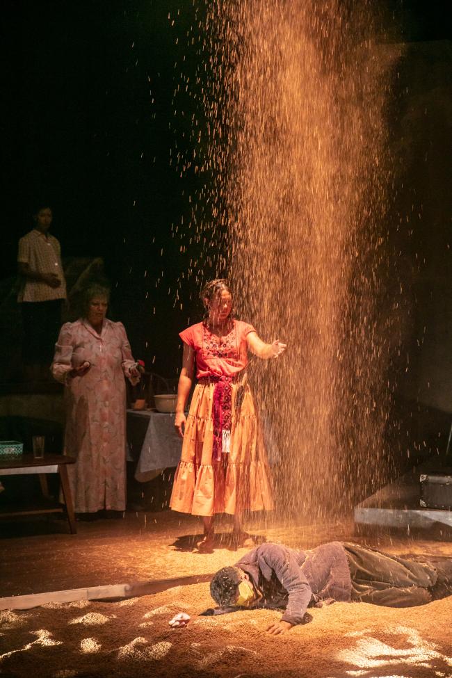 two woman stand on stage with man in far background a light shaft of sand falls from above