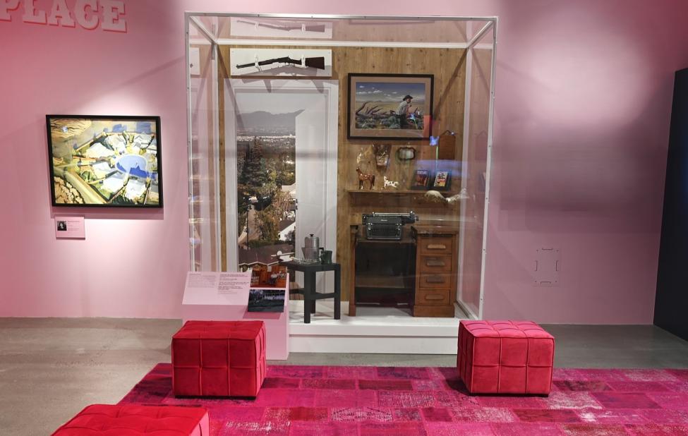 A museum exhibit in a pink room displays a glass case with a typewriter, a table, a lamp, framed photos, and landscape images. Red cushioned seats and a pink-patterned rug are in front of the display.