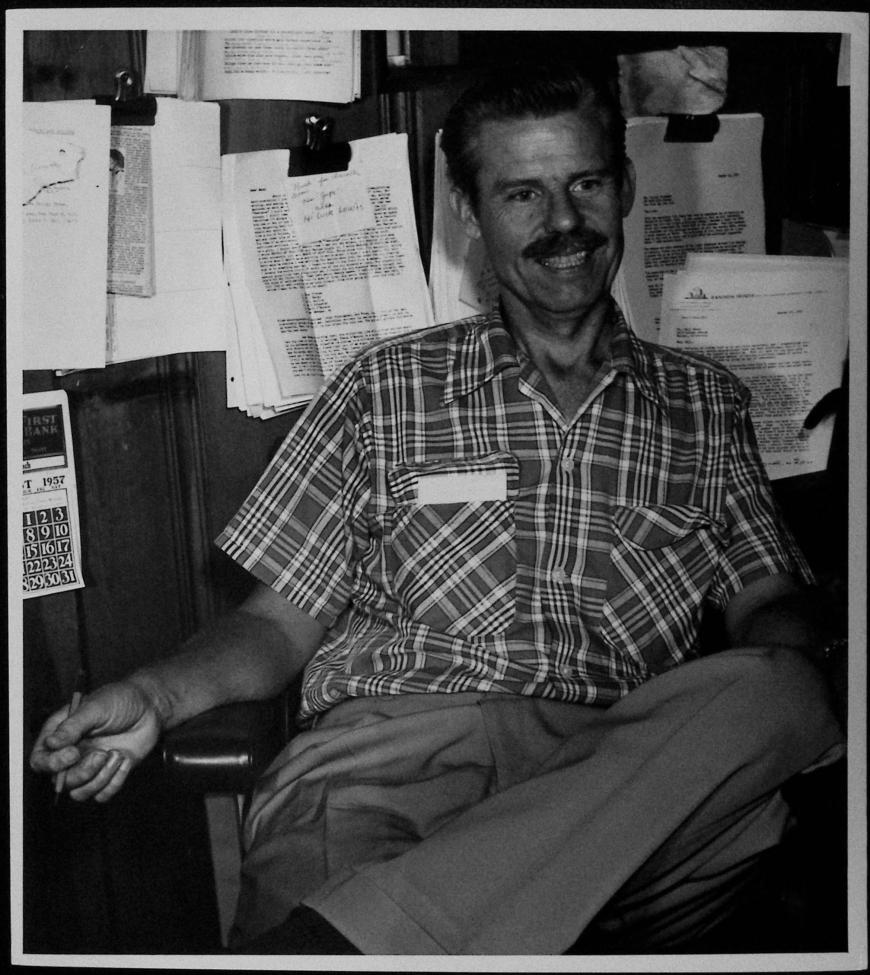 A man with a mustache sits casually, smiling, in an office chair. He wears a plaid shirt and light pants. Behind him, papers and documents are clipped to a wooden wall. His right arm rests casually on the chair arm.