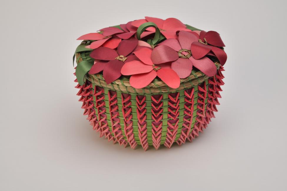 A woven basket with a lid, decorated with pink and red petal-like shapes on top and vertical pink strips around the sides, set against a plain white background.
