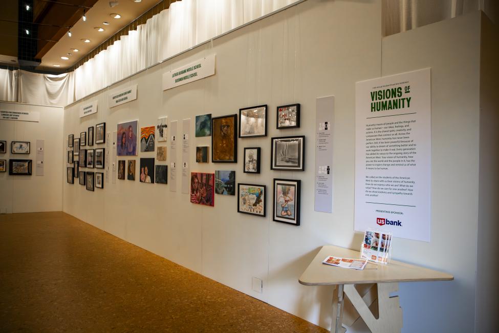 An art gallery wall displays various framed artworks in different styles and sizes, with informational posters including one titled "Visions of Humanity" and a table holding exhibition pamphlets beneath it.