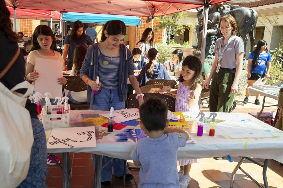 Children and adults gather around a table under a red canopy, engaging in arts and crafts activities with paints and paper at an outdoor event. People chat and work together in a lively, sunny environment.