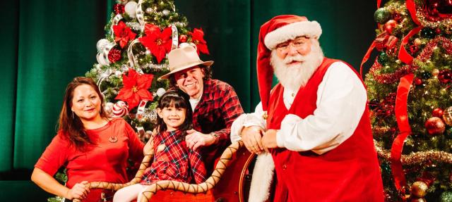 A family poses with Santa Claus in front of decorated Christmas trees. The child sits in a sleigh, smiling, while Santa and her parents stand nearby, all dressed in festive holiday attire.