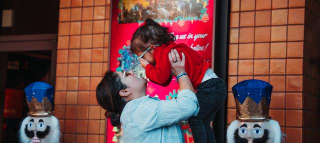 A woman lifts a smiling young girl in a red sweater and glasses, touching noses, in front of a festive display with toy soldier figures and bright red decorations.