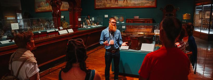 A guide in a denim shirt addresses a small group of people in a dimly lit museum room, surrounded by exhibits and artifacts. A large painting of animals hangs on the wall behind them.