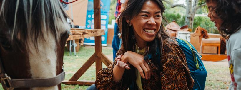 A woman crouches and smiles at a young girl near a horse at an outdoor event, with trees and other people visible in the background.