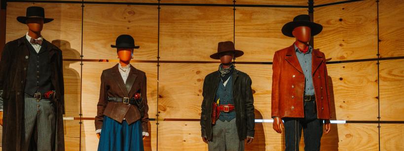 Four mannequins wear classic Wild West costumes, including hats, bowties, vests, long coats, skirts, holsters, and bandanas, standing in front of a wooden panel wall.