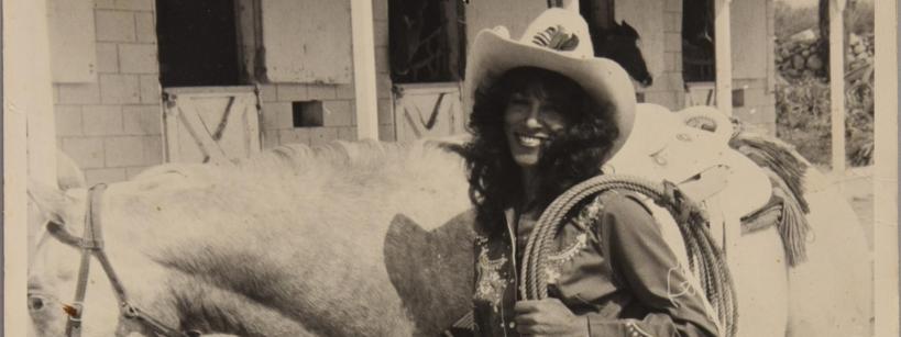 A smiling woman in cowboy attire, including a hat and fringed chaps, stands next to a saddled horse outside a stable, holding a coiled rope. The photo is in black and white.