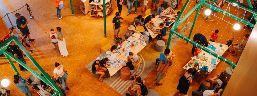 Overhead view of a busy indoor event with people of all ages gathered around tables covered in art supplies and crafts, socializing, and participating in creative activities under bright lighting.