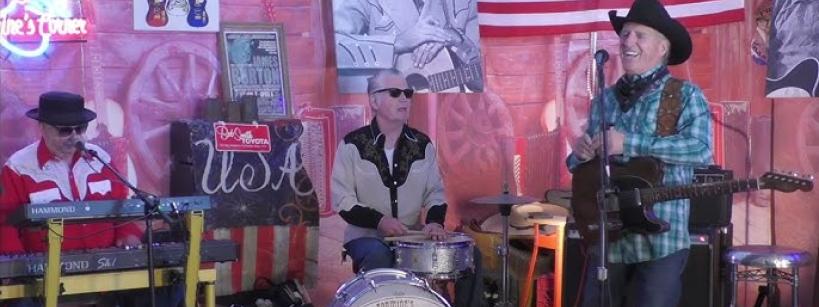 Three musicians perform on stage in a country-themed setting with guitars, American flags, and photos of country legends on the wall. One plays keyboard, one is on drums, and the third plays guitar and sings wearing a cowboy hat.