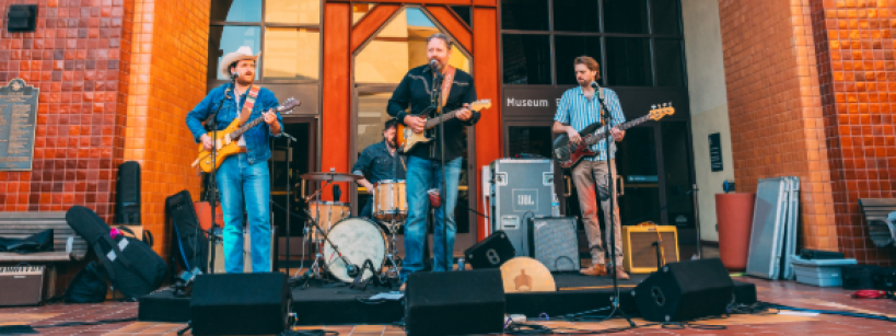 A band performs live music on an outdoor stage beneath the large arched entrance of the Autry Museum, with instruments, speakers, and museum signage visible in the background.