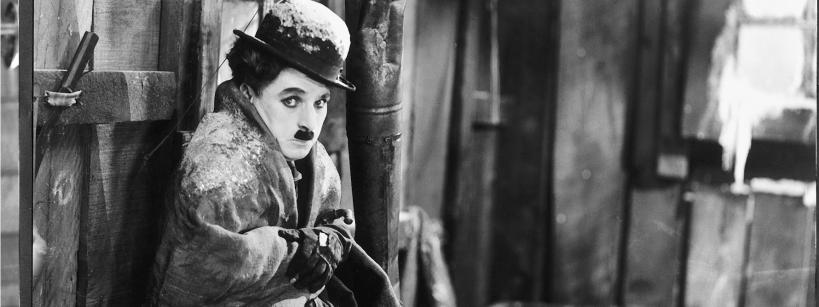 A person dressed in old-fashioned clothes and a bowler hat, with a small mustache, sits shivering in a rustic, snowy cabin, clutching their coat tightly for warmth.