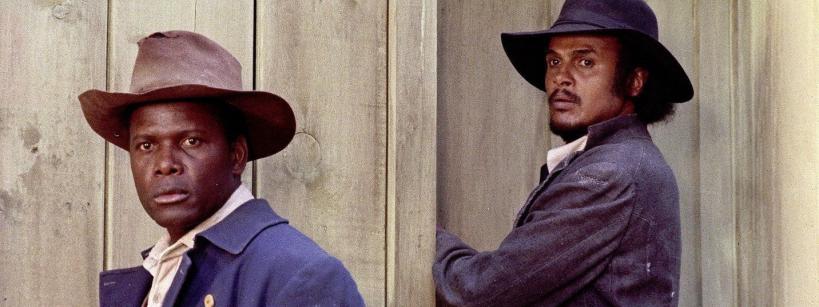 Two men in old-fashioned clothing and wide-brimmed hats stand in front of a wooden wall; one faces forward while the other leans against a door, looking sideways.
