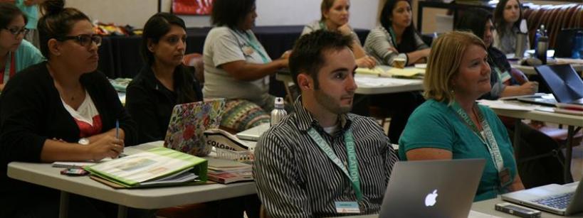 A group of adults sits at tables in a classroom, attentively listening and taking notes. Some have laptops open, and papers and binders are on the tables. The room is well-lit with windows in the background.