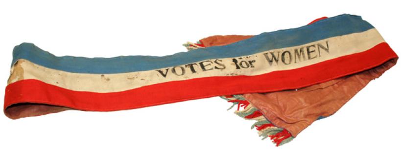 A fabric sash with blue, white, and red stripes, featuring the text "VOTES for WOMEN" printed in black. The ends have multicolored fringe and appear slightly worn.