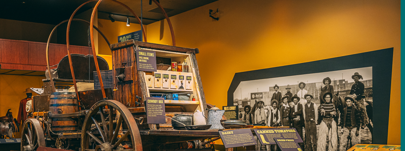 A museum exhibit features a wooden covered wagon stocked with supplies, surrounded by informational signs. Behind it, a large black-and-white photo shows a group of people in period Western clothing.