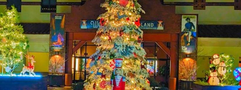 A large, decorated Christmas tree with lights stands in the center of a festive outdoor scene, surrounded by smaller lit displays, including a reindeer, snowmen, and colorful ornaments.