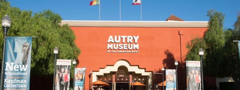 The entrance of the Autry Museum of the American West, with orange walls, three flags flying above, banners on either side, and people near the entrance on a sunny day.