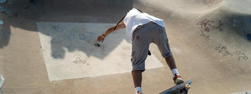 A skateboarder in a white shirt and gray shorts prepares to drop into a concrete skatepark bowl covered in graffiti, balancing on the edge with knees bent and arms outstretched.