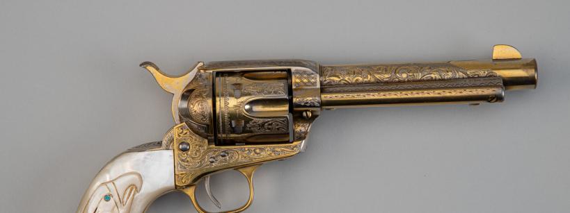 A decorative revolver with an engraved gold-colored barrel and cylinder, and a white handle featuring an eagle design and a blue gemstone eye, displayed on a plain gray background.