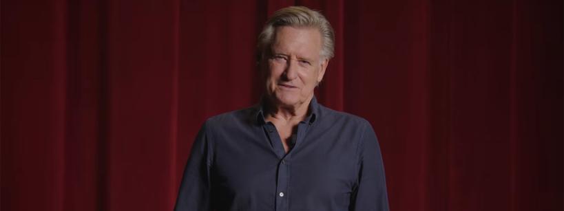 A man with gray hair wearing a navy shirt stands in front of red theater curtains, looking at the camera and smiling gently.