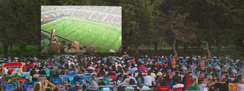 A large outdoor crowd sits on lawn chairs facing a screen showing a soccer match in a stadium. Trees line the background, creating a park-like setting.