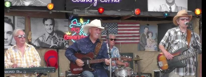 Four men perform country music onstage with guitars, keyboard, and drums. They wear casual clothes and hats. Neon signs, American flags, and black-and-white portraits decorate the background.