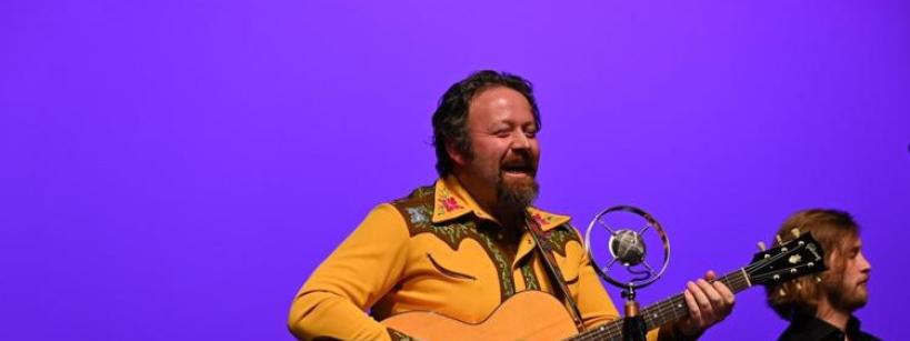 A man in a yellow shirt plays an acoustic guitar and sings into a microphone on stage with a purple background. Another musician stands beside him.