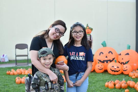 A woman and two children smile at a pumpkin patch decorated with large, inflatable jack-o’-lanterns. The boy wears a camouflage outfit and holds a small pumpkin, while the girl wears a navy shirt and glasses.